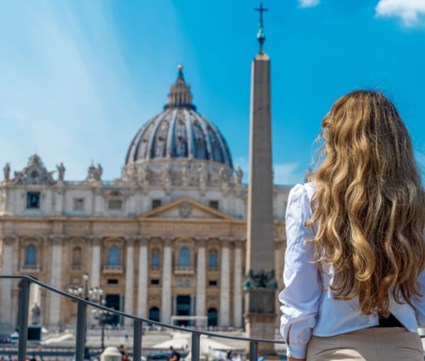 View of the blonde woman from behind in Vatican City. Saint Peter Square with multiple tourists, Saint Peter Basilica and the Vatican Obelisk on the background